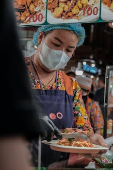 Street food vendor wearing mask and apron serves delicious local cuisine in a busy market setting.