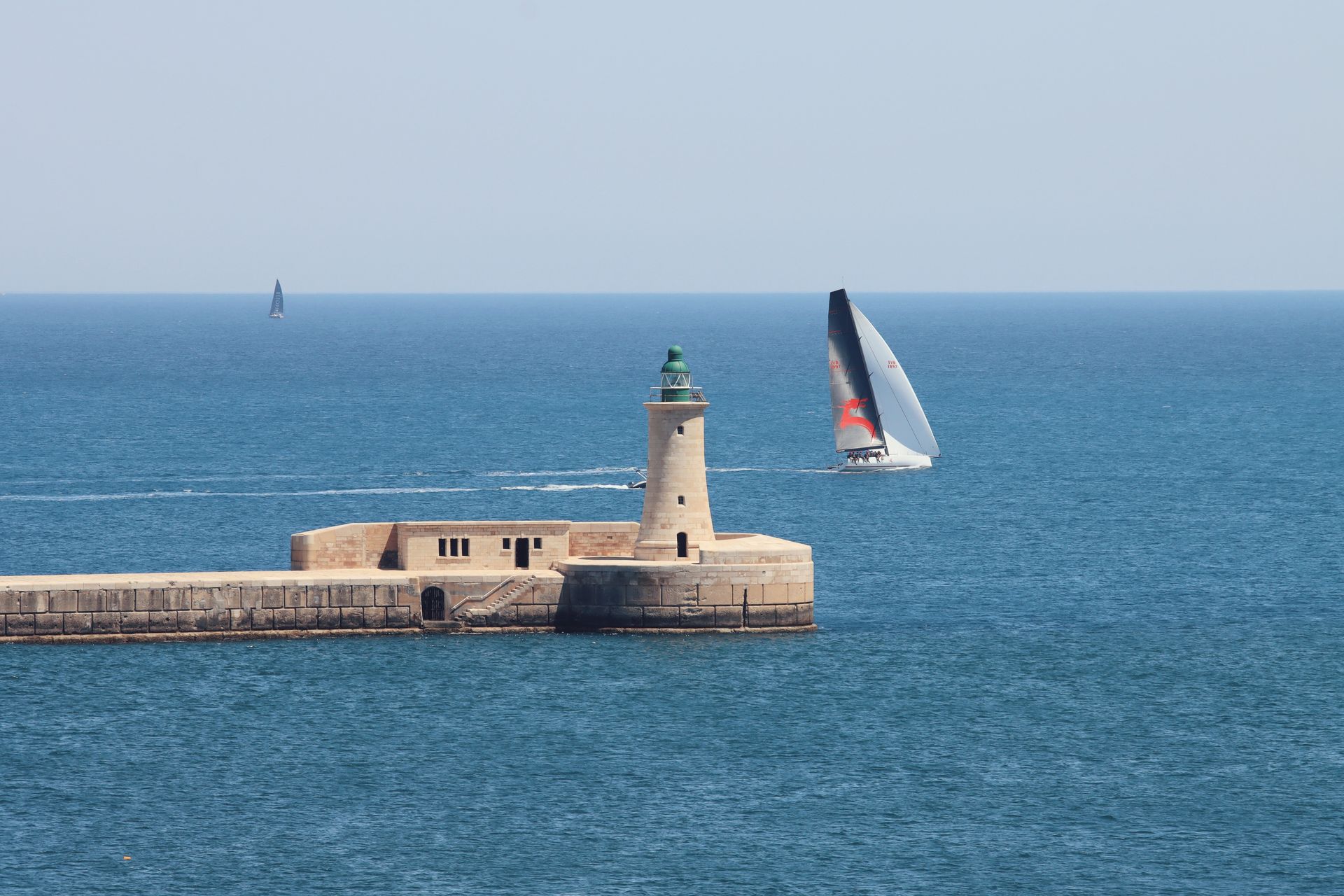 Lighthouse at Valletta harbor with sailboats on a clear blue sea. Iconic Mediterranean seascape.