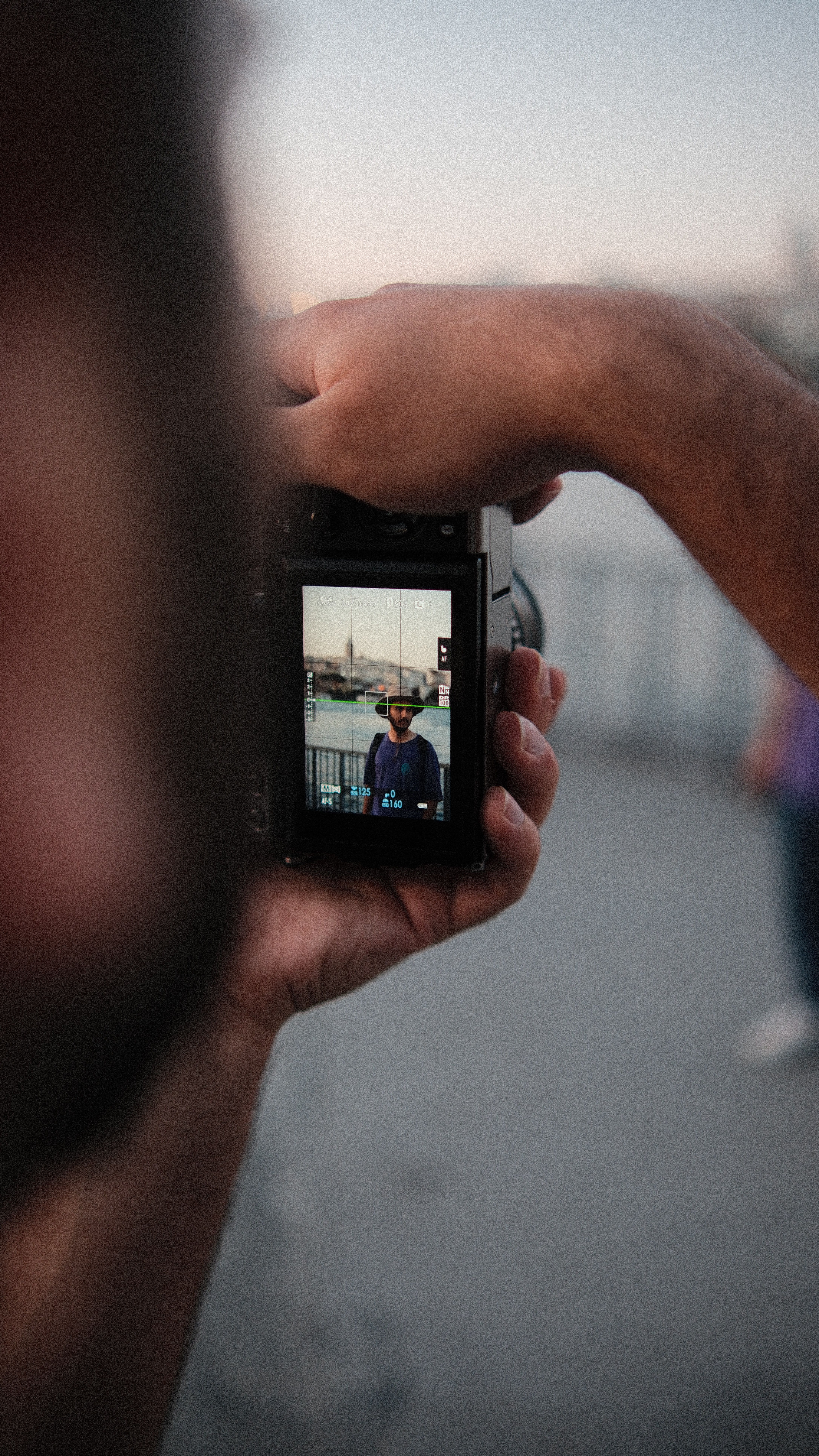 Close-up of hands holding a camera capturing a man's portrait by the waterfront.