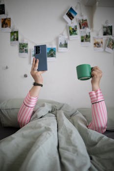 Woman in striped pajamas holds a phone and cup in bed, surrounded by photos.