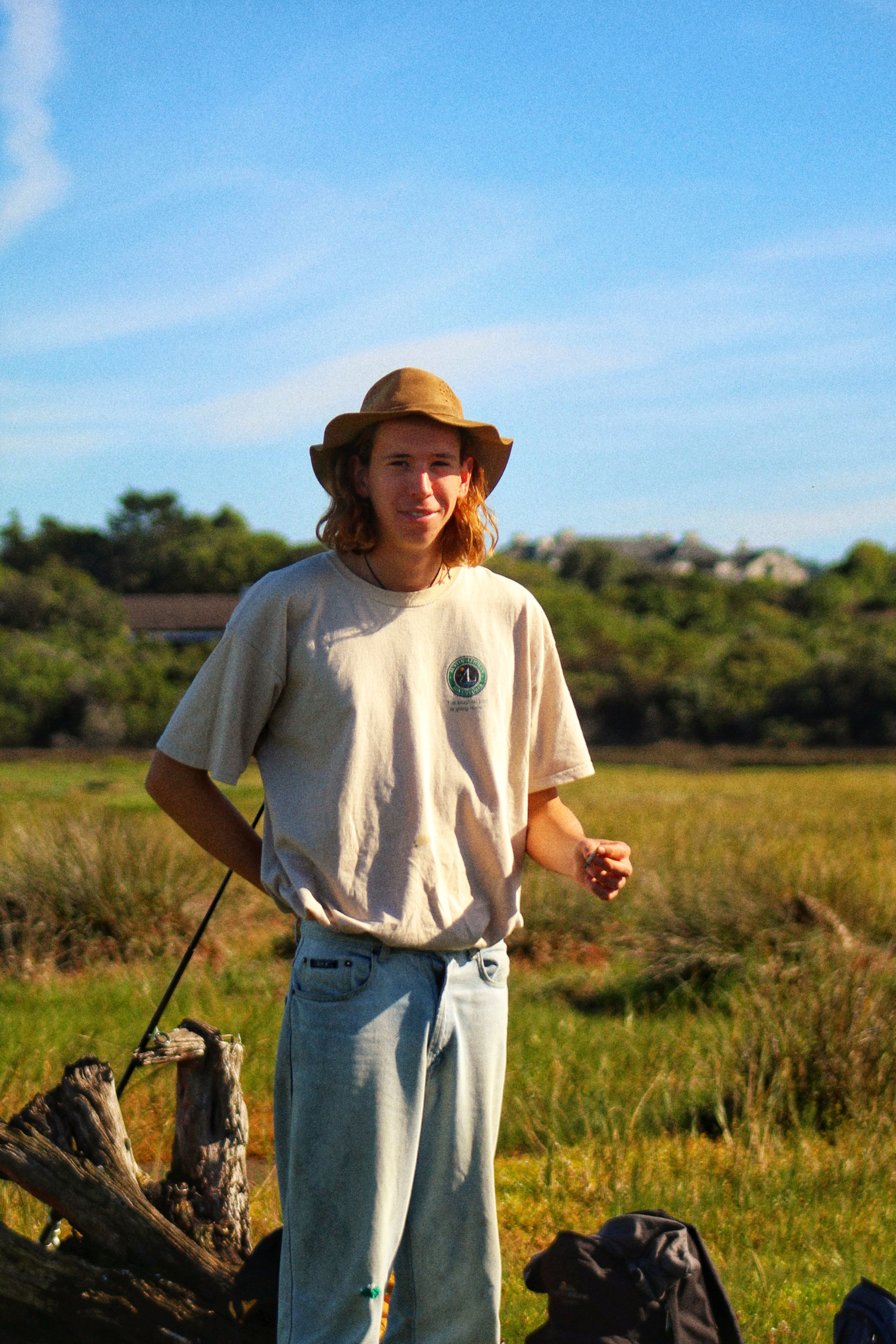 A man with long hair and a hat stands in a sunny field, surrounded by grass and trees.