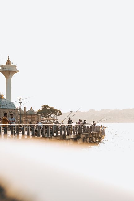People fishing on a scenic pier during a bright, sunny day with a lighthouse in the background.