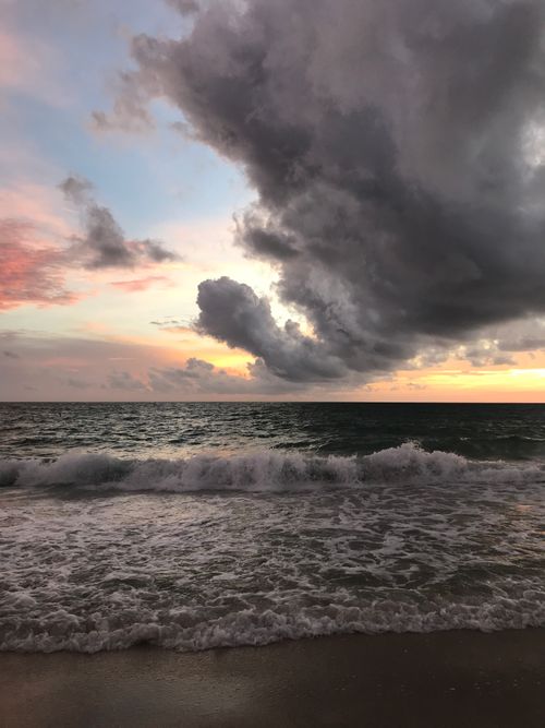 Free A striking beach scene at sunset with dramatic storm clouds and waves crashing on the shore. Stock Photo