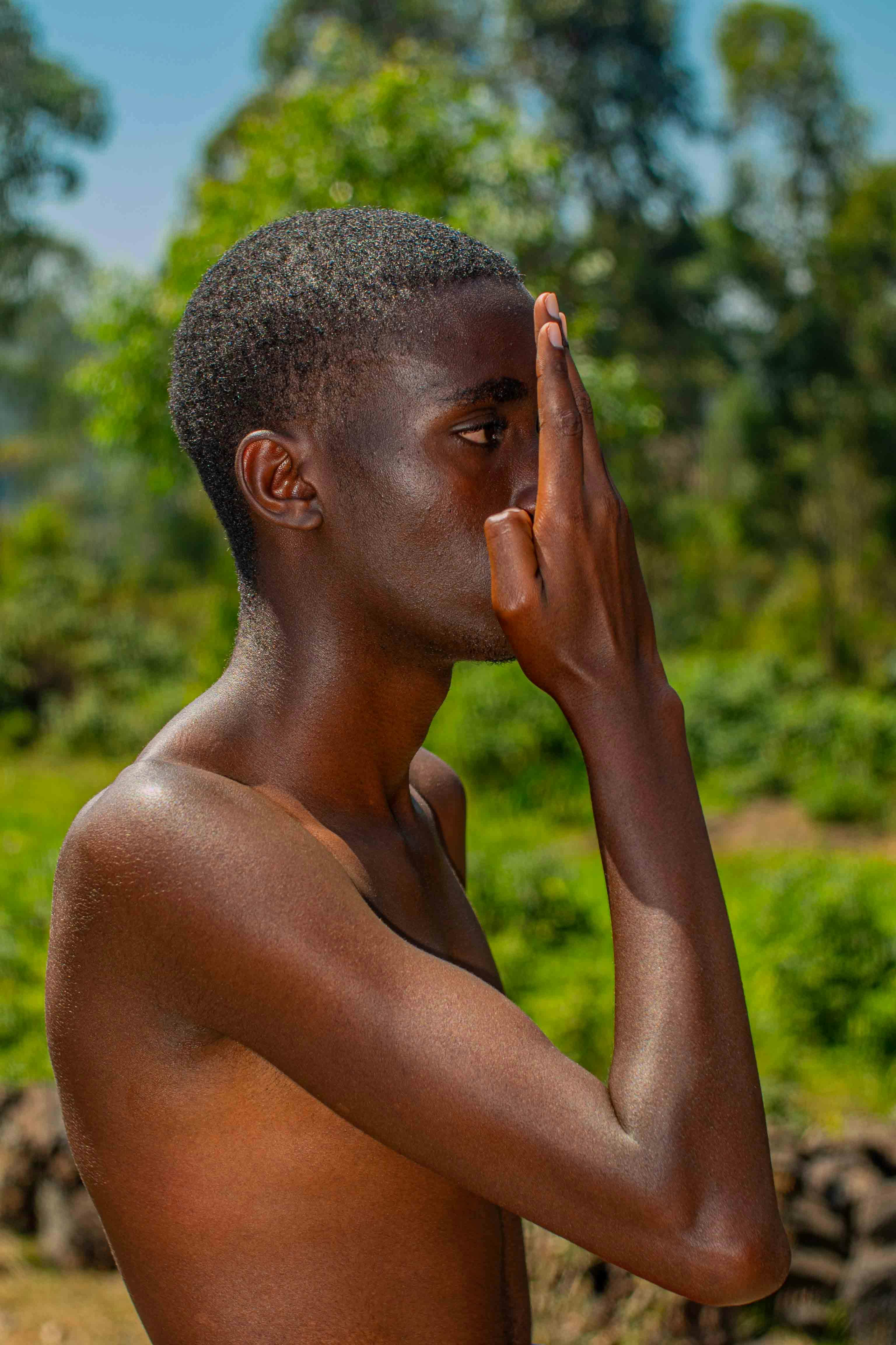 Shirtless man standing outdoors, covering face with hand, in a lush green setting.