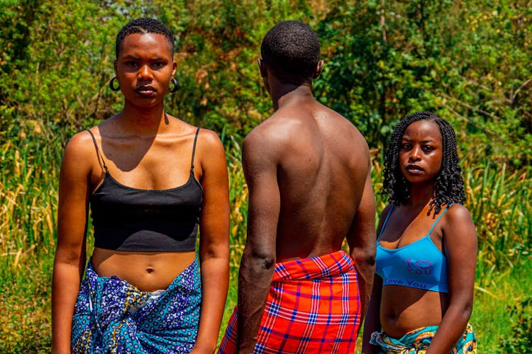 Young Women And A Man Standing On A Field In Casual Summer Outfits 