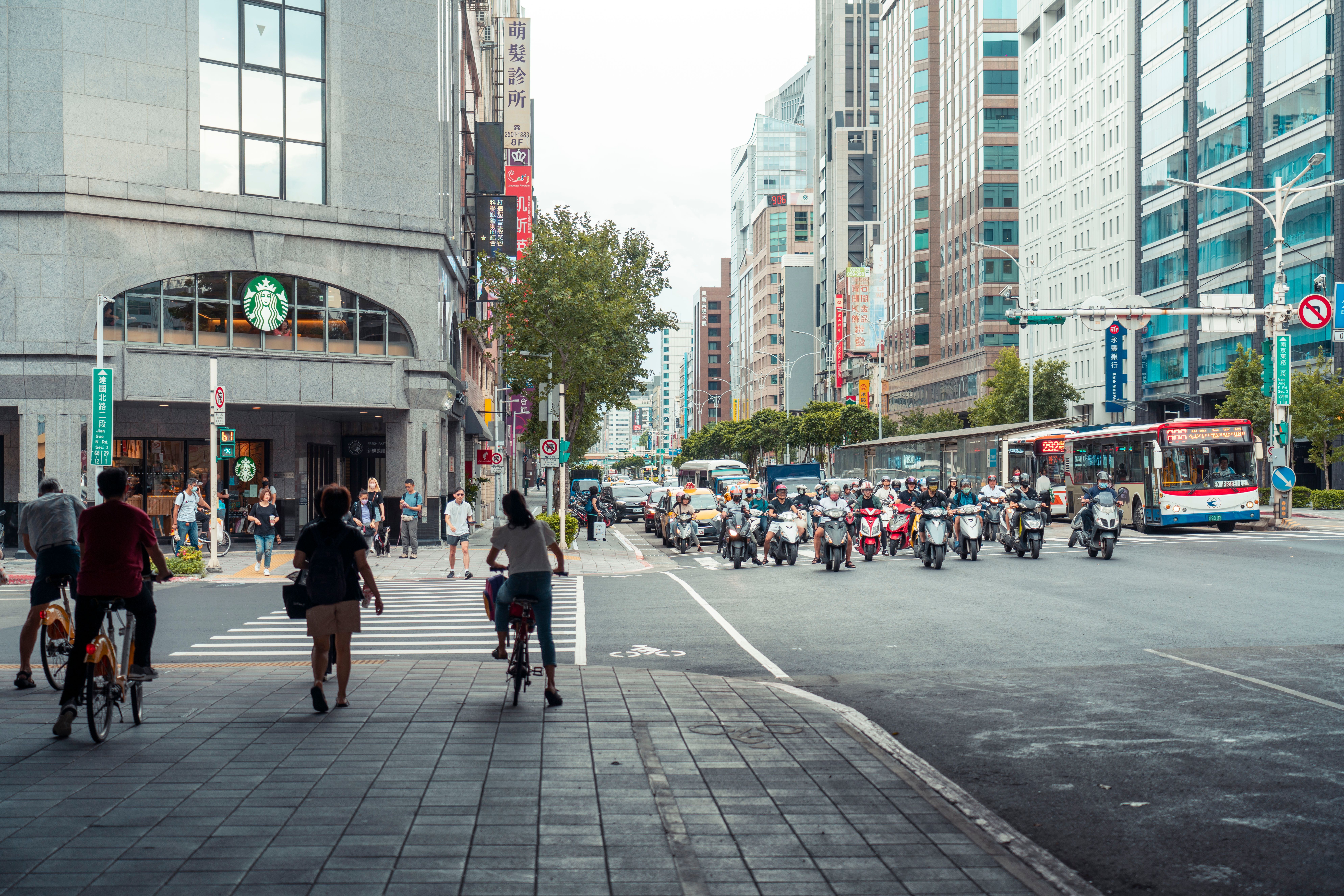 Busy city street scene showing bikers, scooters, and pedestrians in a bustling downtown area.