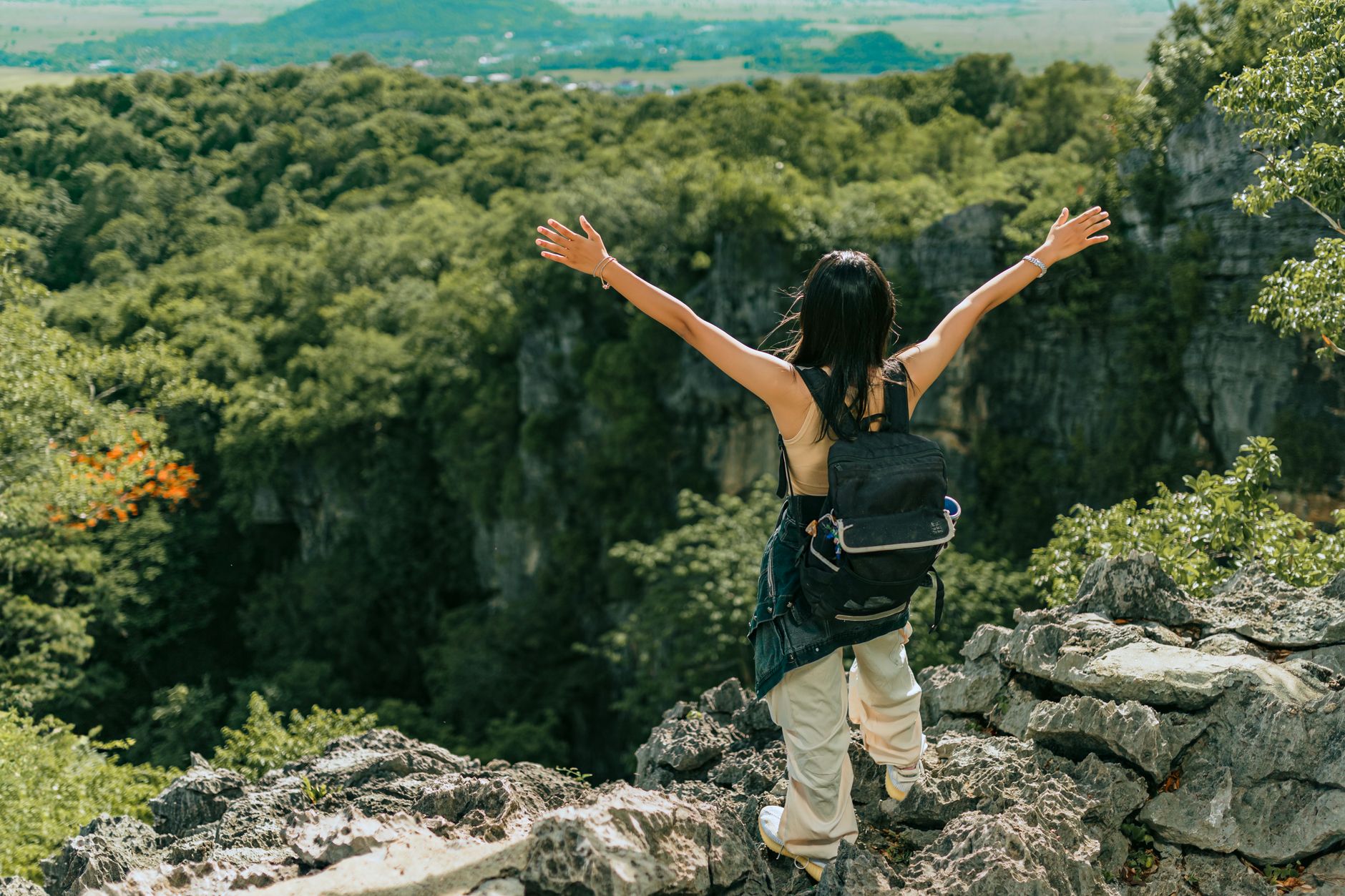 Back view of a woman enjoying a scenic mountain landscape, arms raised in joy, surrounded by lush green forest.