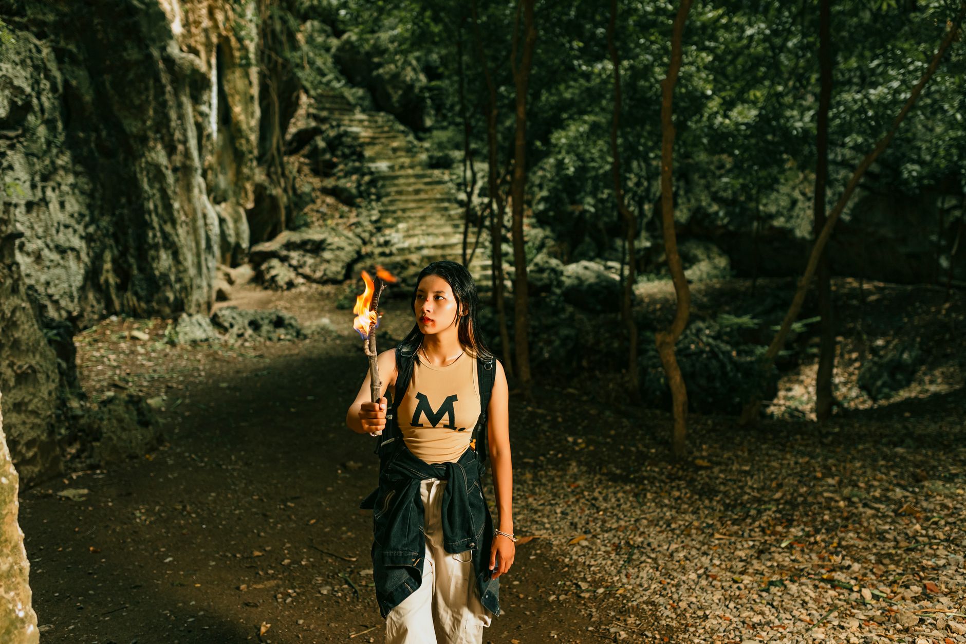 A woman explores a cave with a torch in a forest setting, highlighting adventure and exploration.