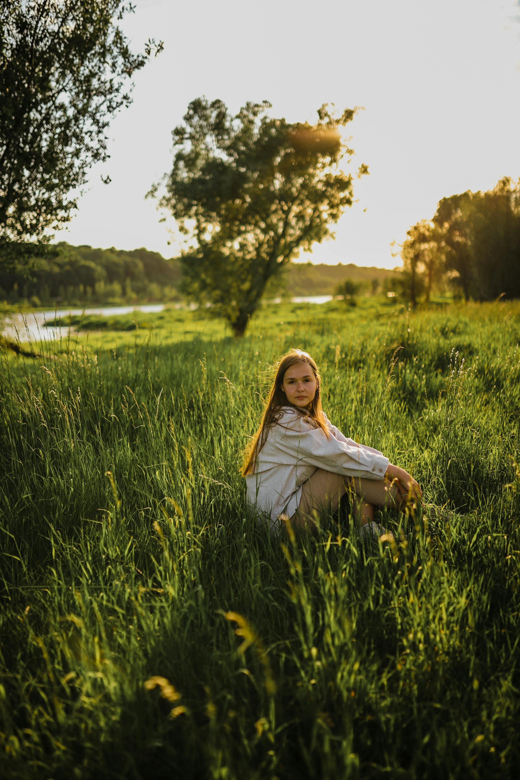 A young woman sitting peacefully in a lush green meadow during sunset, exuding tranquility and natural beauty.