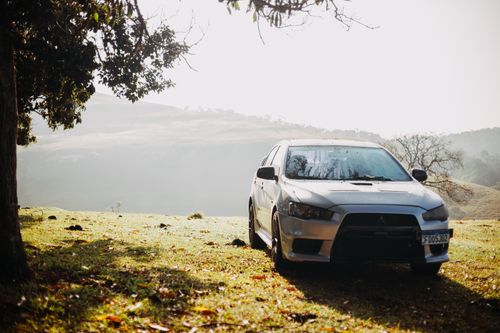 Free A silver car parked in a sunny rural meadow with a picturesque hilly landscape and trees. Stock Photo