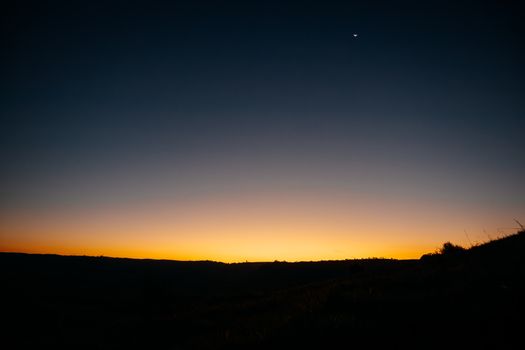 A tranquil sunset with a clear sky over silhouette of rolling plains, featuring a distant crescent moon.