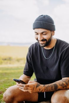 A man in a black t-shirt and knit hat using a smartphone outdoors, smiling.