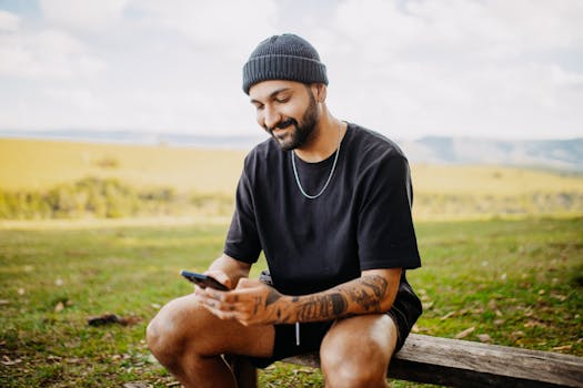 Mid adult man sitting on a bench outdoors, using a smartphone, wearing casual clothing and a hat.