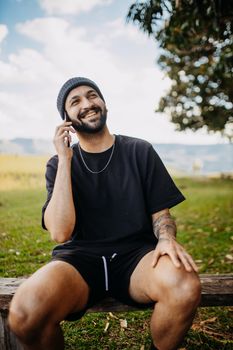 A smiling man in casual wear sits on a bench outdoors, talking on his smartphone.