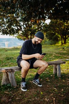 Casual young man sitting outdoors, smiling while using smartphone on a wooden bench.