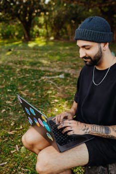 Casually dressed man with tattoos using a laptop in a grassy outdoor setting.
