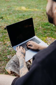 Tattooed man sitting on a log using a laptop in a green meadow.