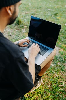 A person typing on a laptop while sitting on the grass in a rural meadow area.