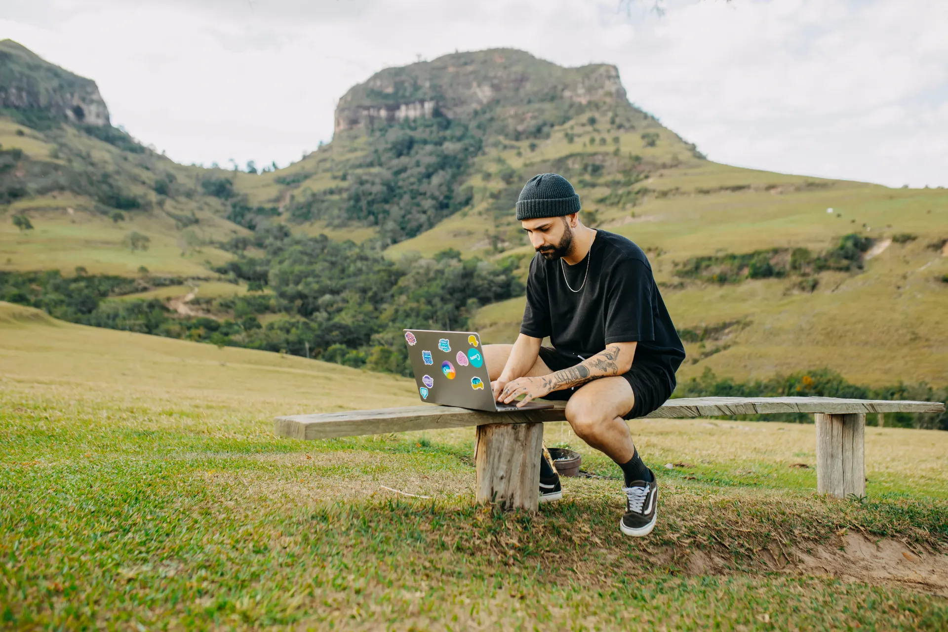 A man working on a laptop on a wooden bench in front of rocky hills. A man working on a laptop on a wooden bench in front of rocky hills.