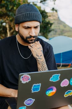 A man sits outdoors contemplating work on a sticker-covered laptop with a nature backdrop.