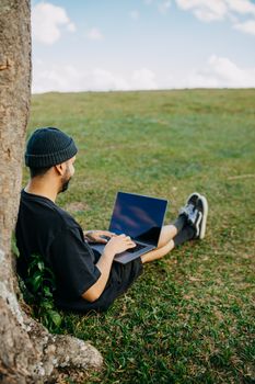 Man sitting against a tree, working on a laptop in a grassy field. Perfect for outdoor work themes. - Fotografer Sidoarjo