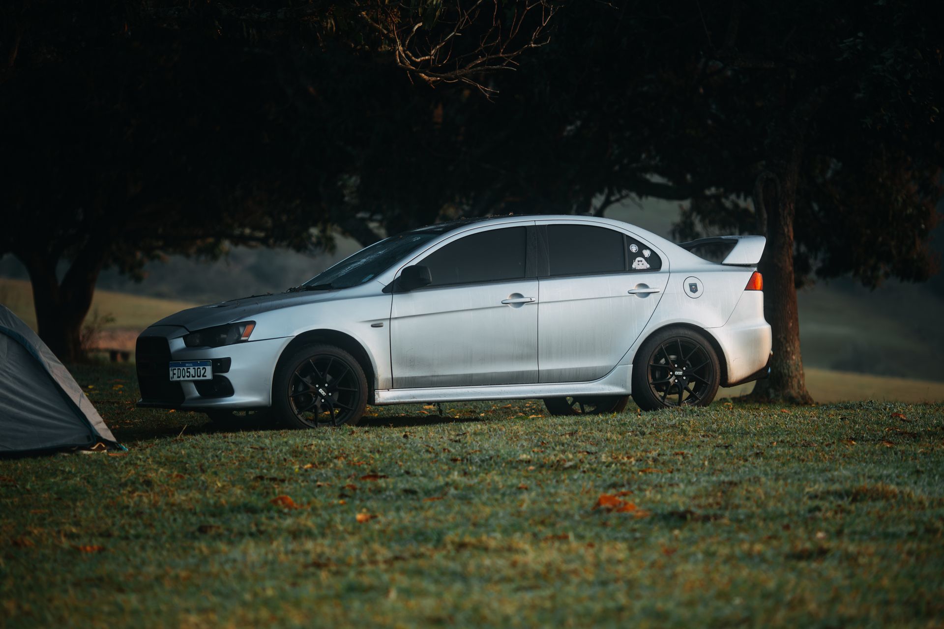 A sleek silver Mitsubishi Lancer Evolution parked outdoors on grass with a scenic backdrop.