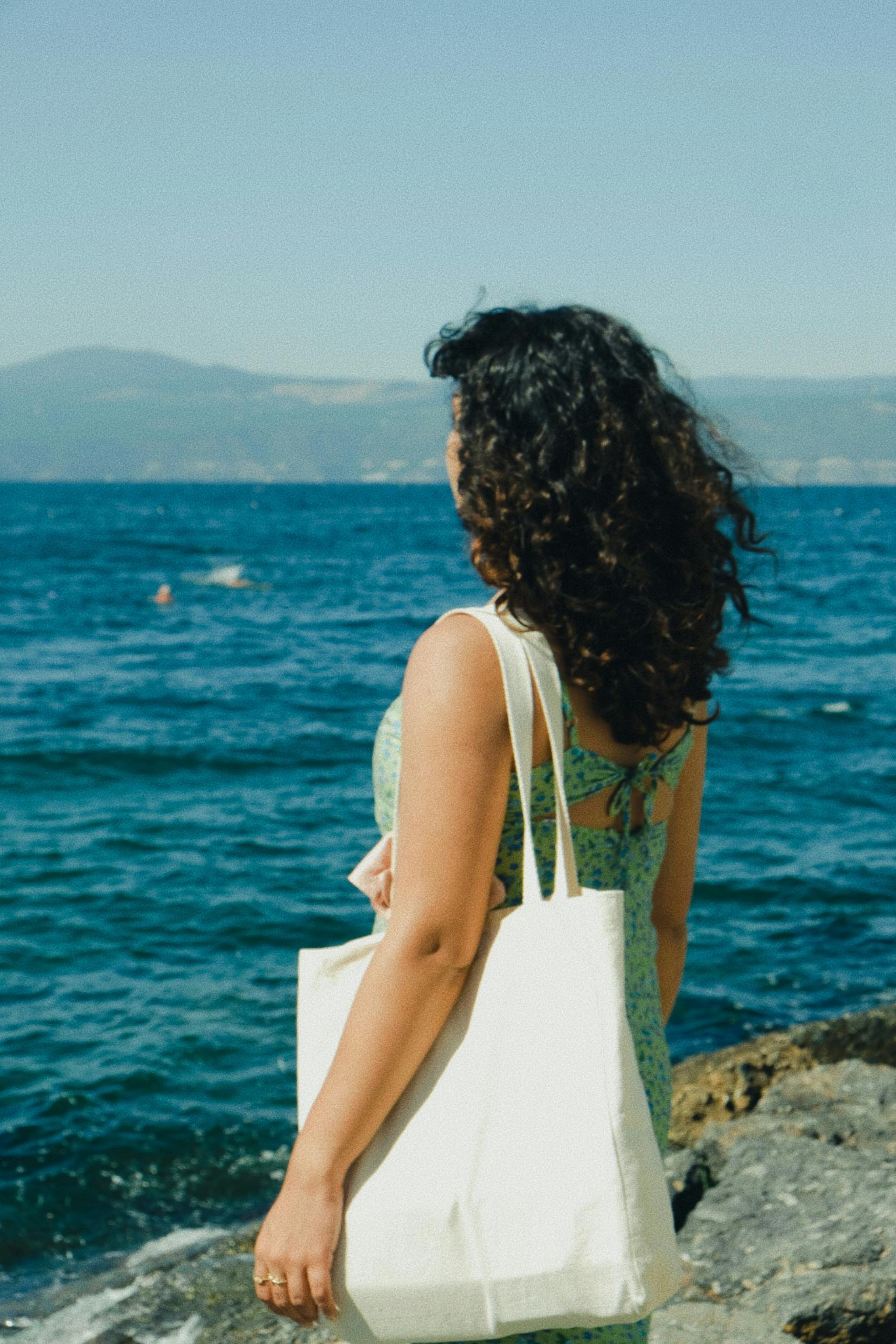 Brunette woman in a summer dress stands by the sea, enjoying the coastal view with a tote bag.