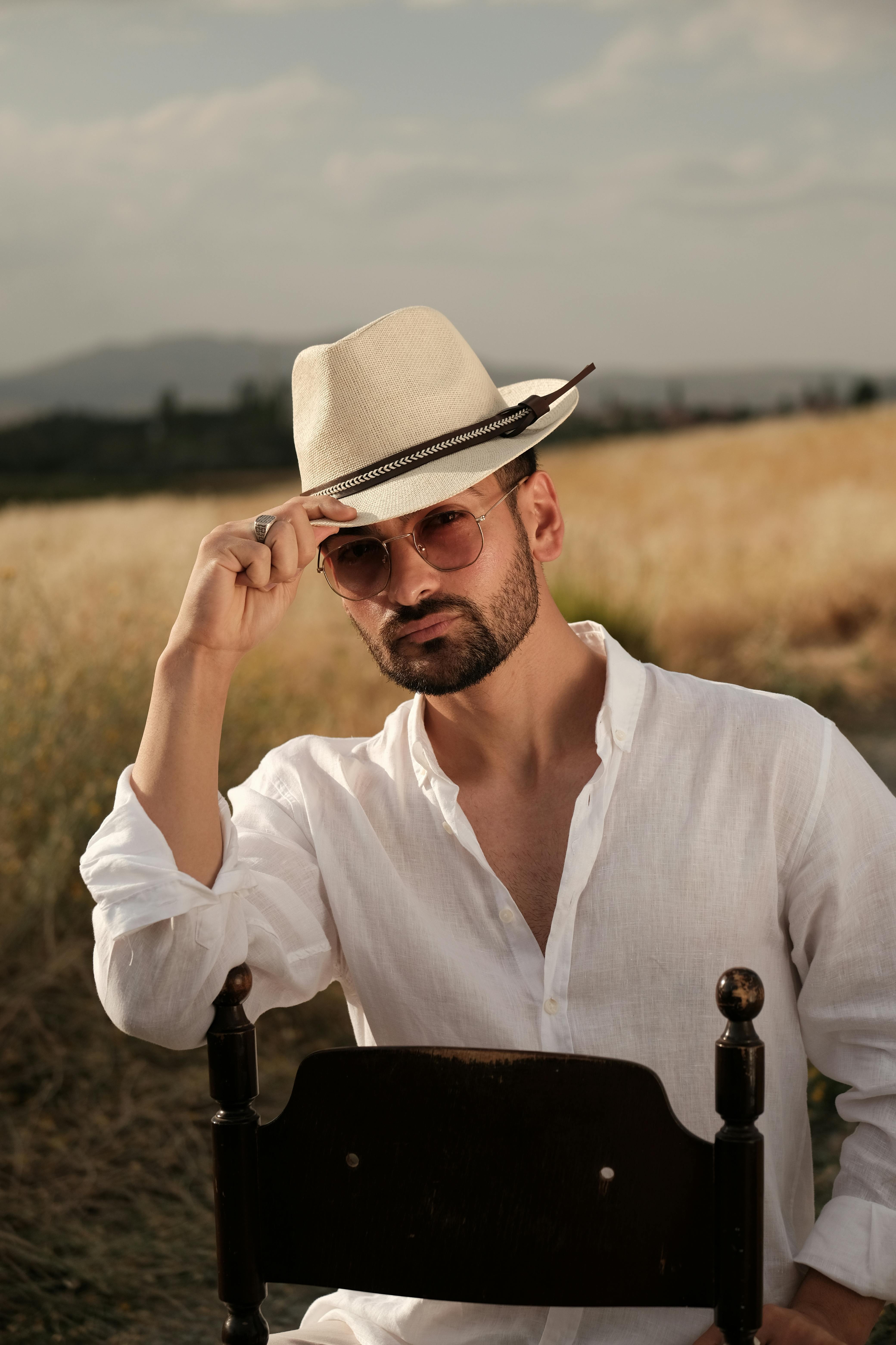 A fashionable man in sunglasses and hat sitting outdoors in a sunny rural setting.