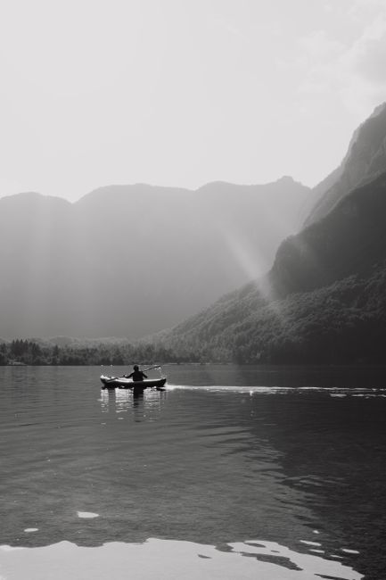 A solitary kayaker on a tranquil lake surrounded by majestic mountains in black and white.
