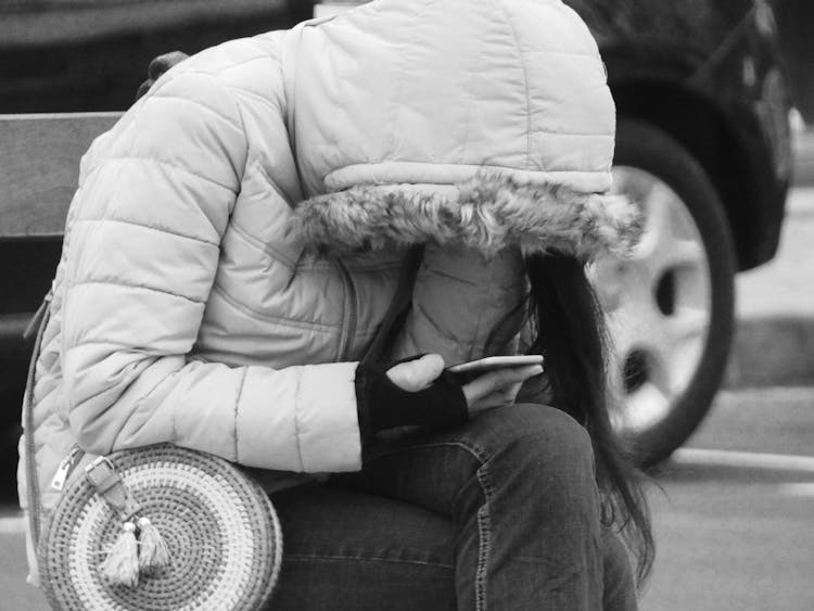 Woman In A Jacket With A Hood Sitting On A Street And Looking At Her Phone 