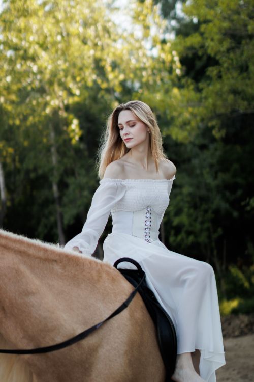 Free A serene scene of a woman in a white dress horseback riding through lush greenery. Stock Photo