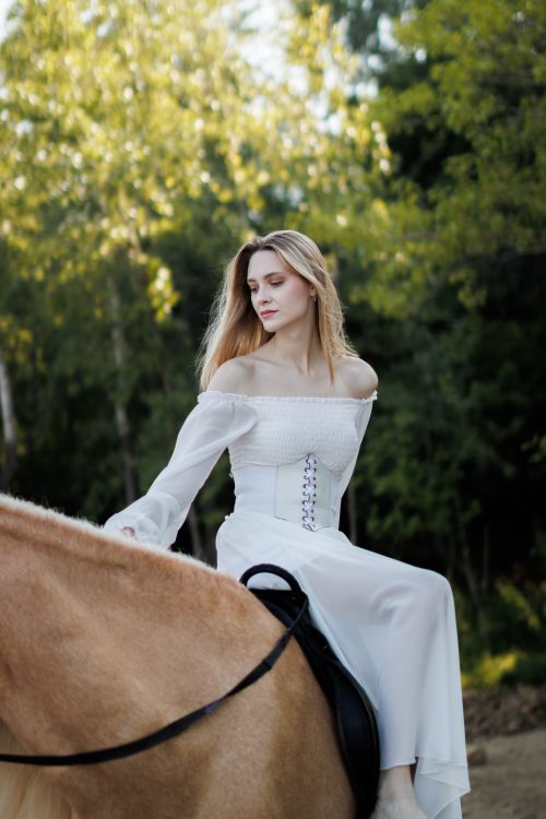 Free A woman in a long-sleeved white dress riding a horse in a wooded area, showcasing elegant fashion and nature. Stock Photo