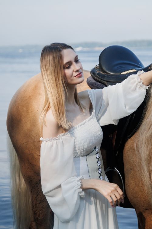 Free Serene scene of a blonde woman in a white dress posing with a horse near water. Stock Photo
