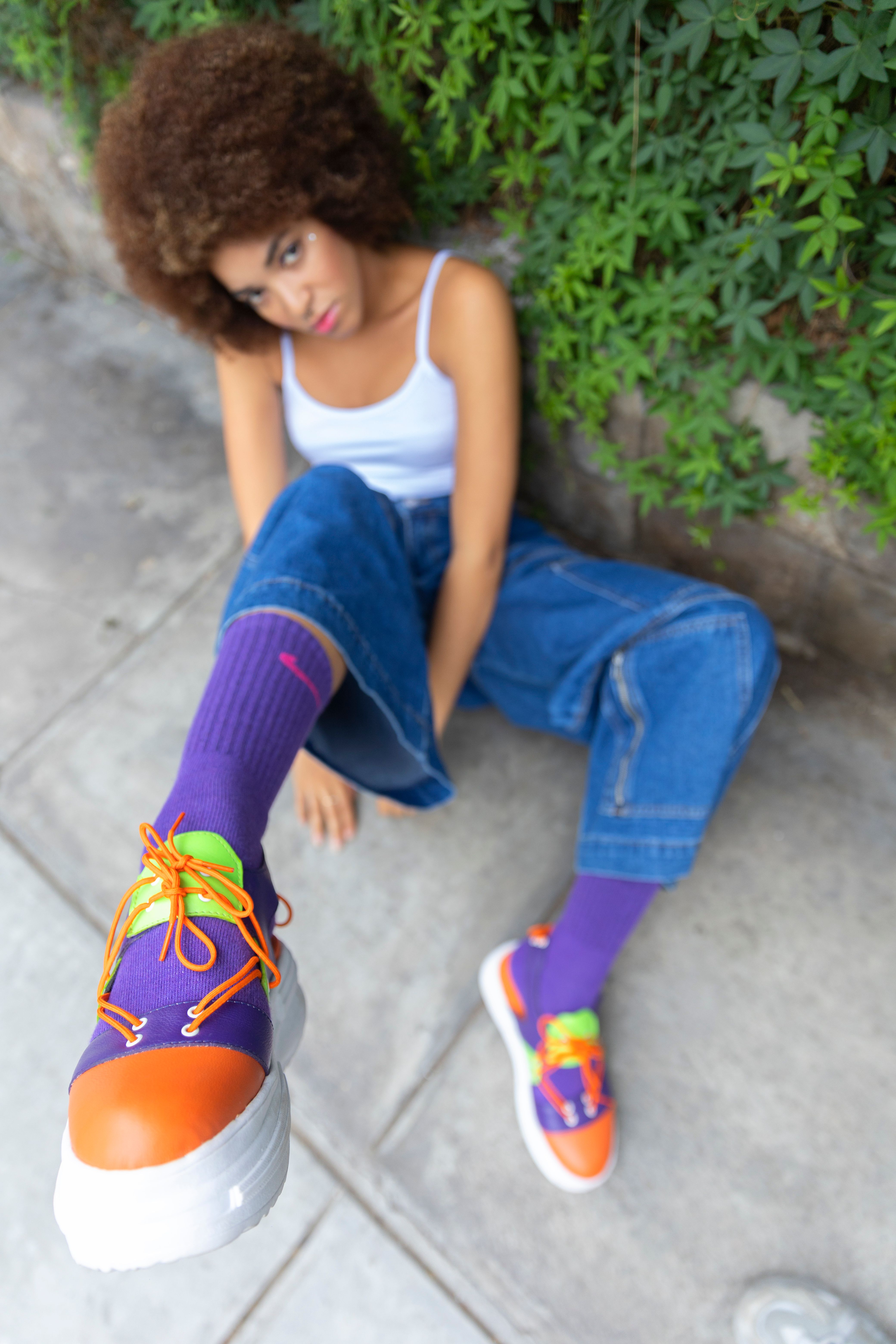A young woman outdoors in vibrant sneakers and casual fashion, sitting against a green backdrop.