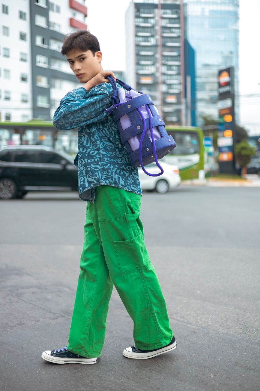 Model in green pants and patterned shirt posing on a city street with a stylish bag.