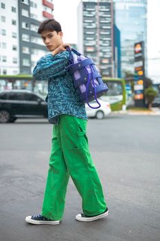 Model in green pants and patterned shirt posing on a city street with a stylish bag.