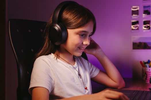 Young girl smiling while using a laptop and wearing headphones in a cozy indoor setting.