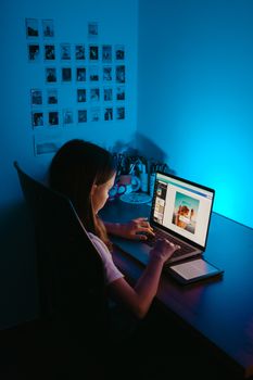 Teen girl working on a laptop under blue light in a dimly lit room, focusing on digital projects.