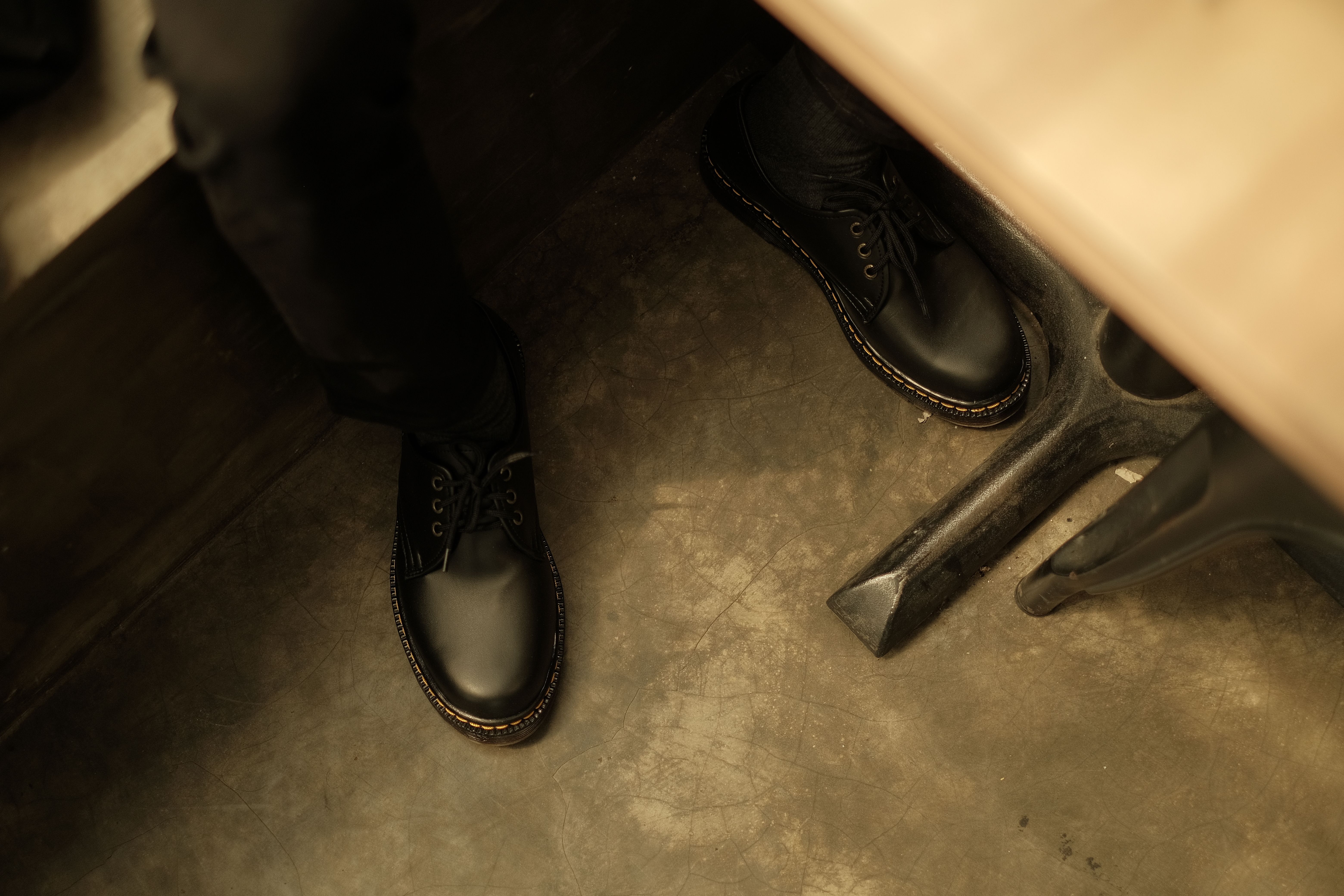 Black leather boots seen under a table against a concrete floor, indoor setting.