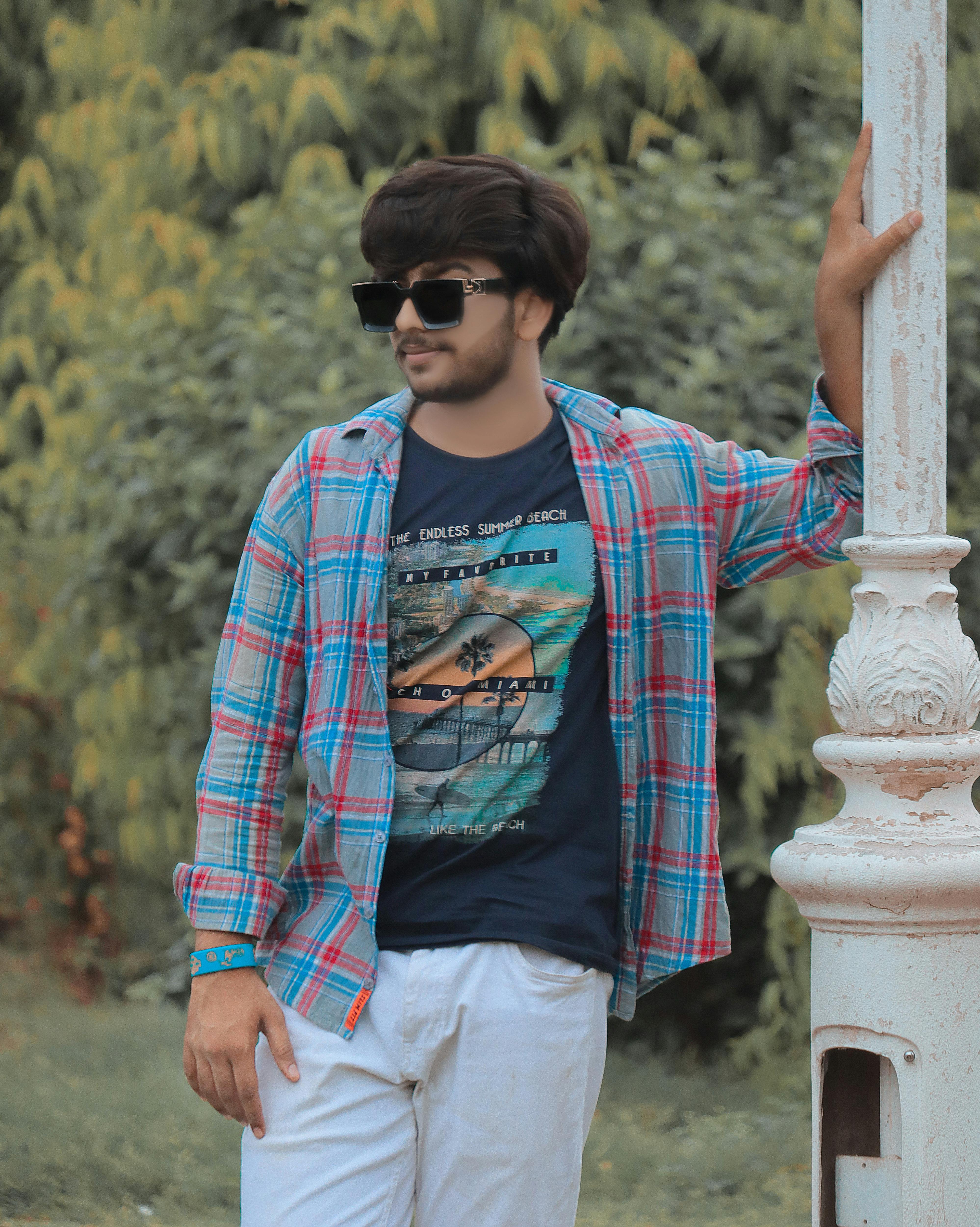 Stylish young man wearing sunglasses and checked shirt posing in a park during the day.