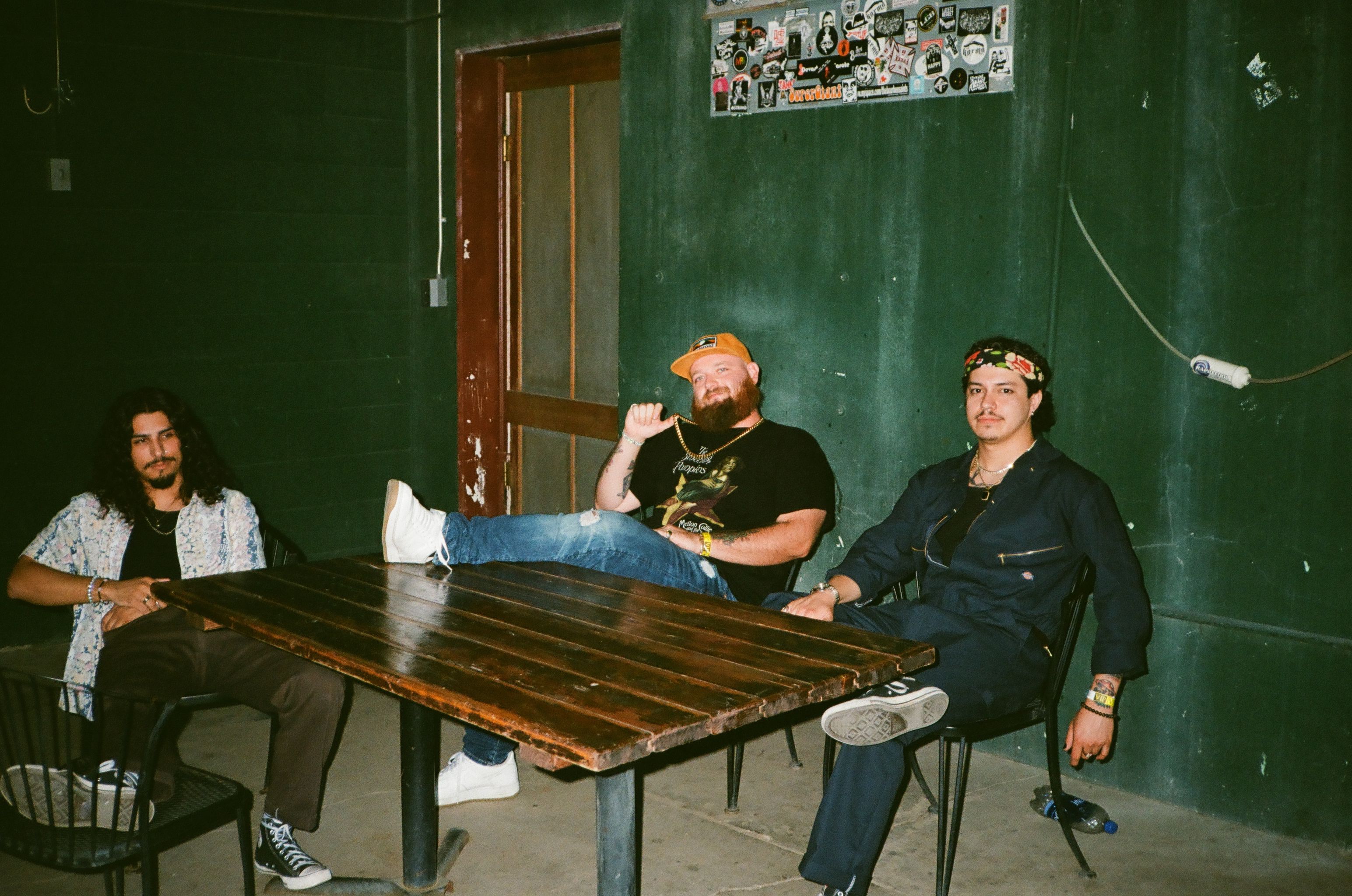 Three men sit casually around a wooden table in an urban setting, displaying a relaxed and rebellious vibe.