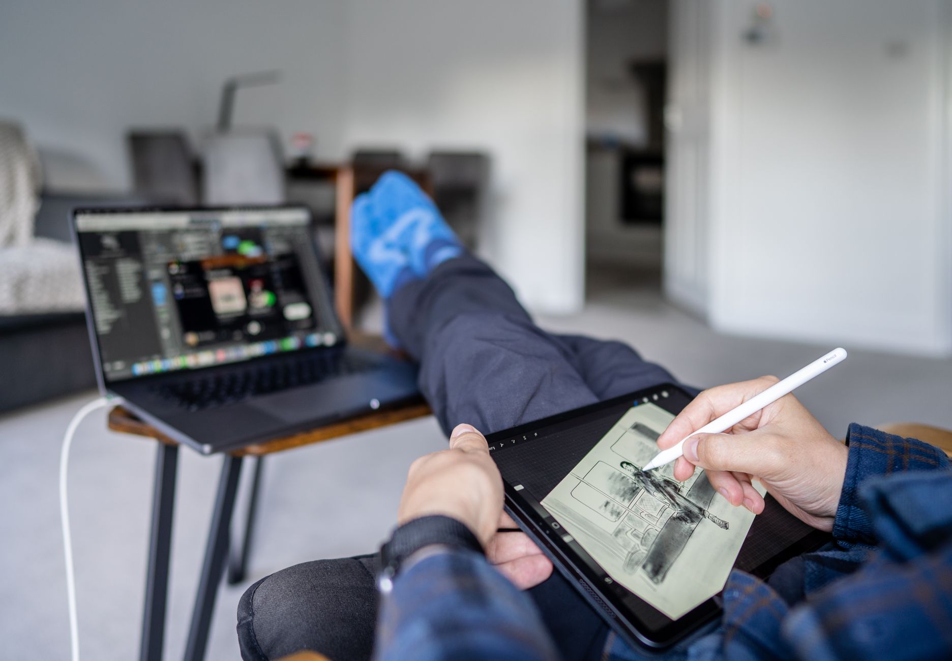 Person creating digital art on tablet while multitasking with laptop in a cozy indoor setting.