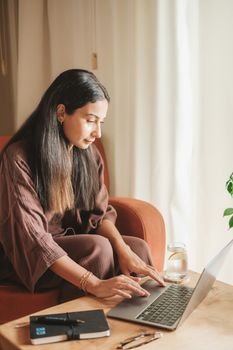 Woman with long hair working on a laptop at home, sitting comfortably in natural light.