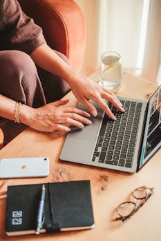 A woman using a laptop on a wooden table with a phone, notepad, and glasses nearby.