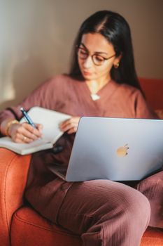 A woman sitting on a chair writing notes in a notebook while using a laptop.