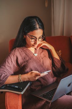 Young woman multitasking on phone and laptop while sitting in a cozy armchair indoors.
