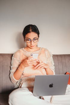 A focused woman with glasses using smartphone and laptop, sitting comfortably indoors.