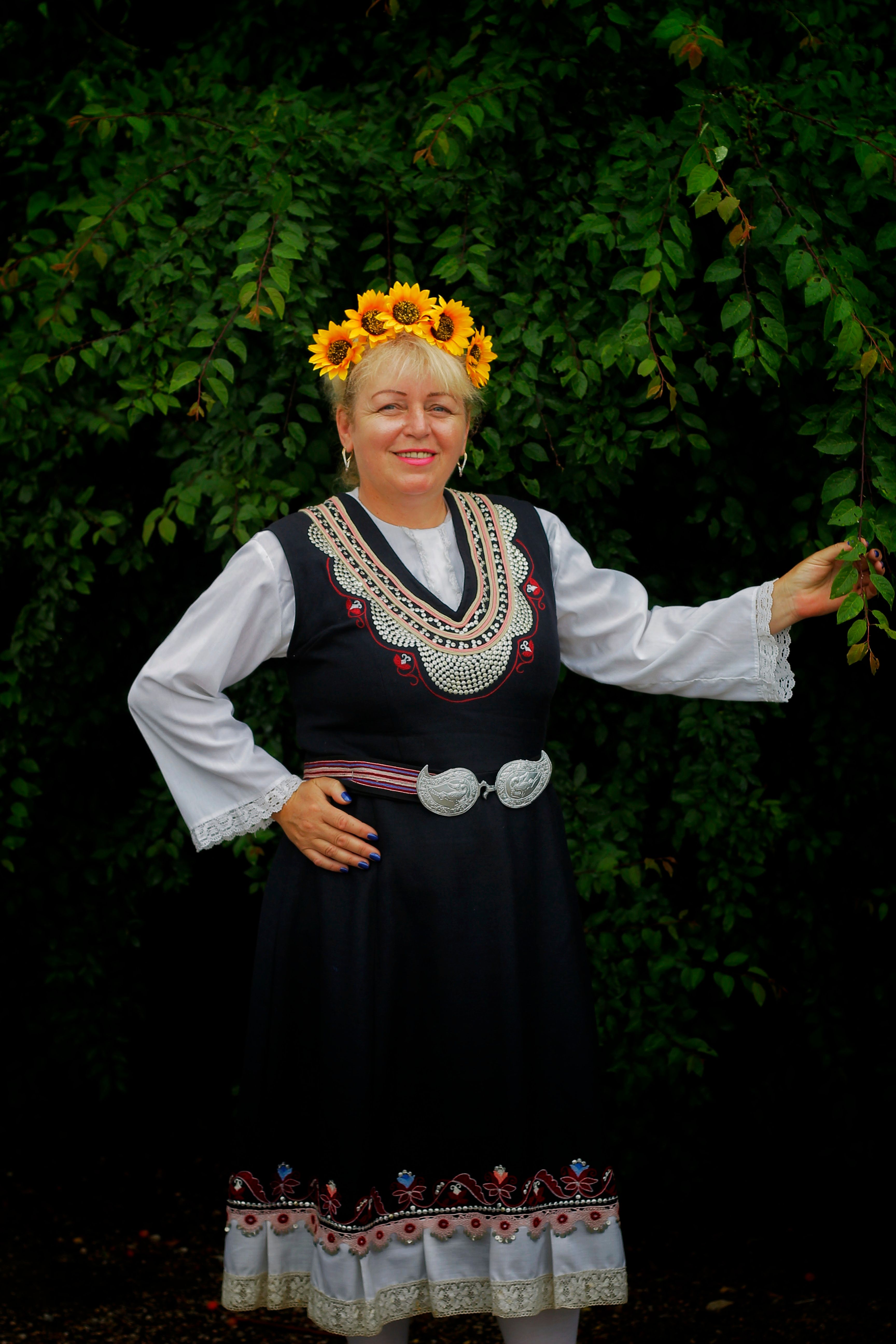 Smiling woman in Balkan traditional attire with sunflower crown outdoors.