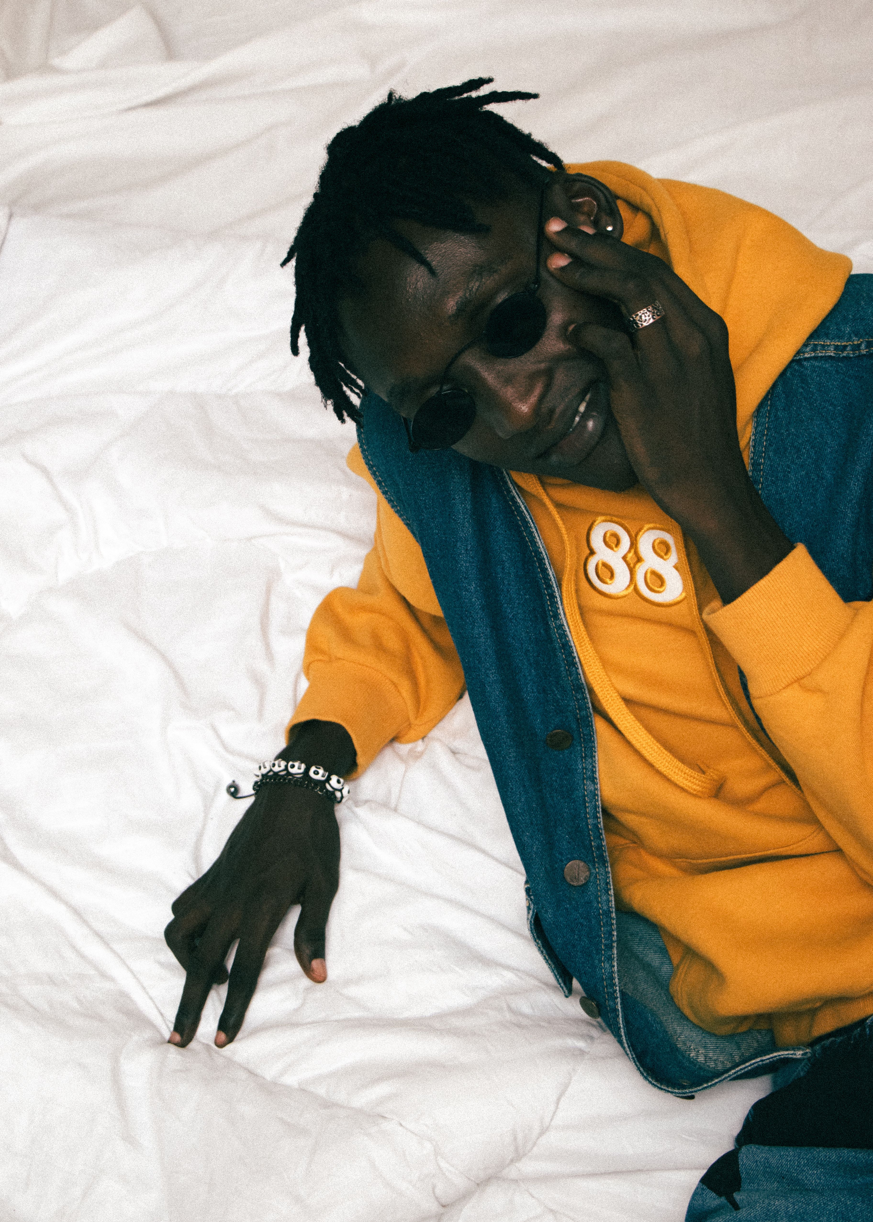 Fashionable young man wearing a yellow hoodie, sunglasses, and jewelry, posing on a bed.