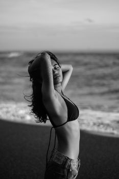 A serene black and white portrait of a woman standing on the beach in a bikini with her arms raised.